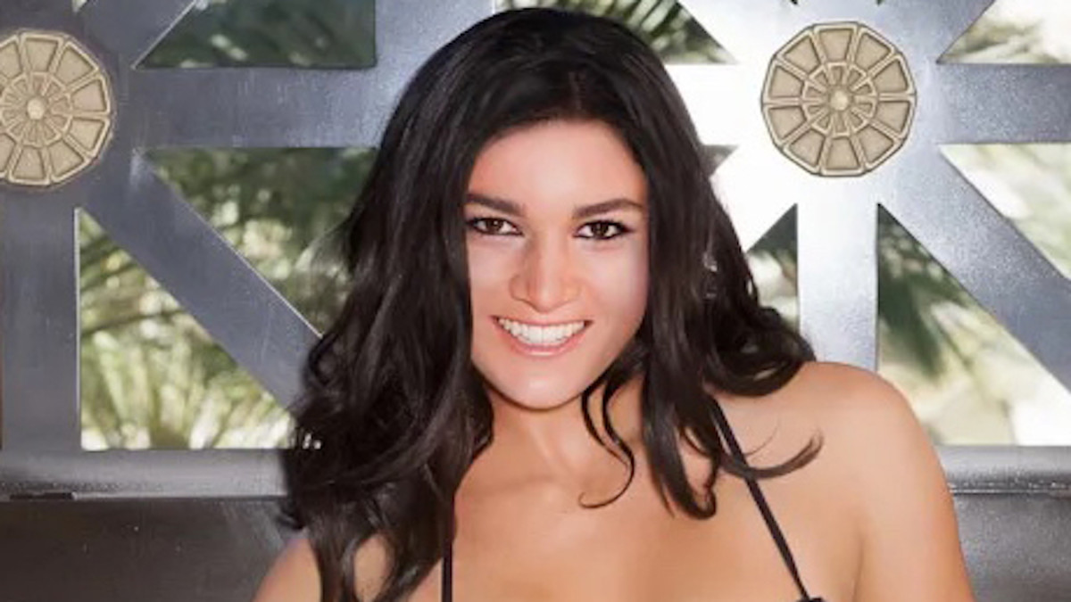 Michelle Jenneke poses in a dark purple string bikini in front of a wooden and glass-paneled wall.